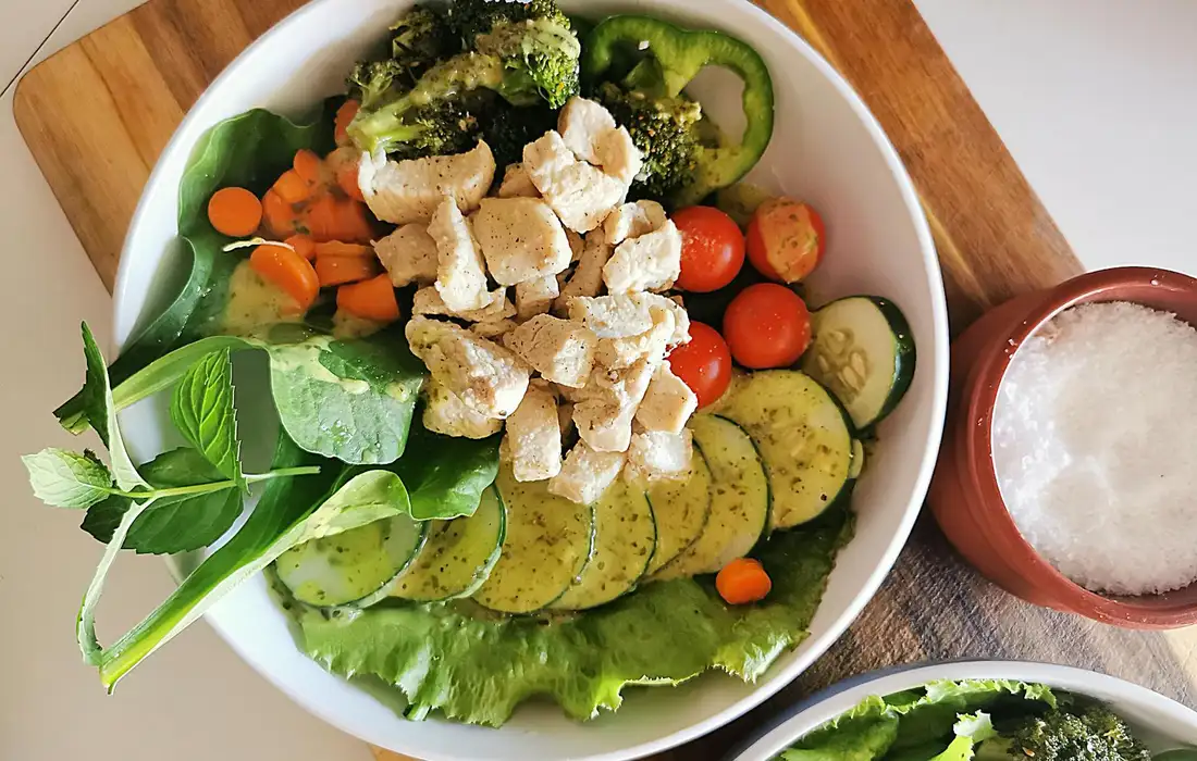 A white bowl of mixed greens, broccoli florets, cucumber slices, carrot rounds, cherry tomatoes, and diced chicken on top, with a salt shaker nearby.