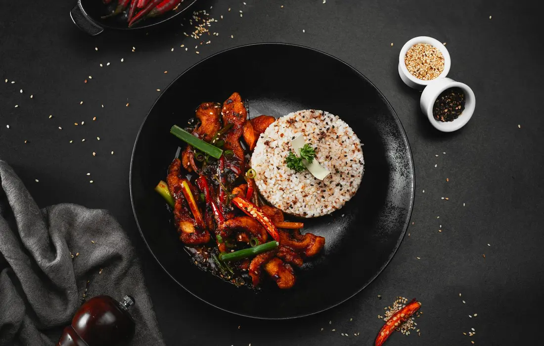 Plate of grits with roasted peppers and greens on a dark background, with two small bowls of seasoning nearby.