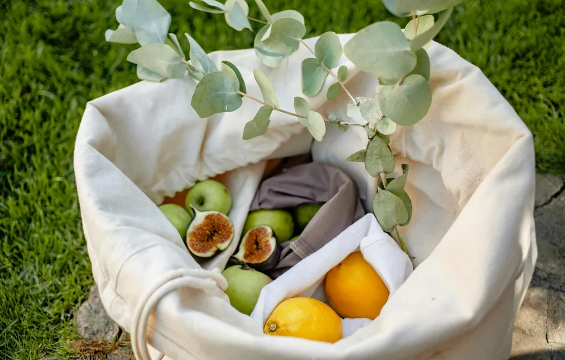 A white cloth tote bag filled with fresh green apples, a few other fruits, and sprigs of eucalyptus, resting on a grassy surface.