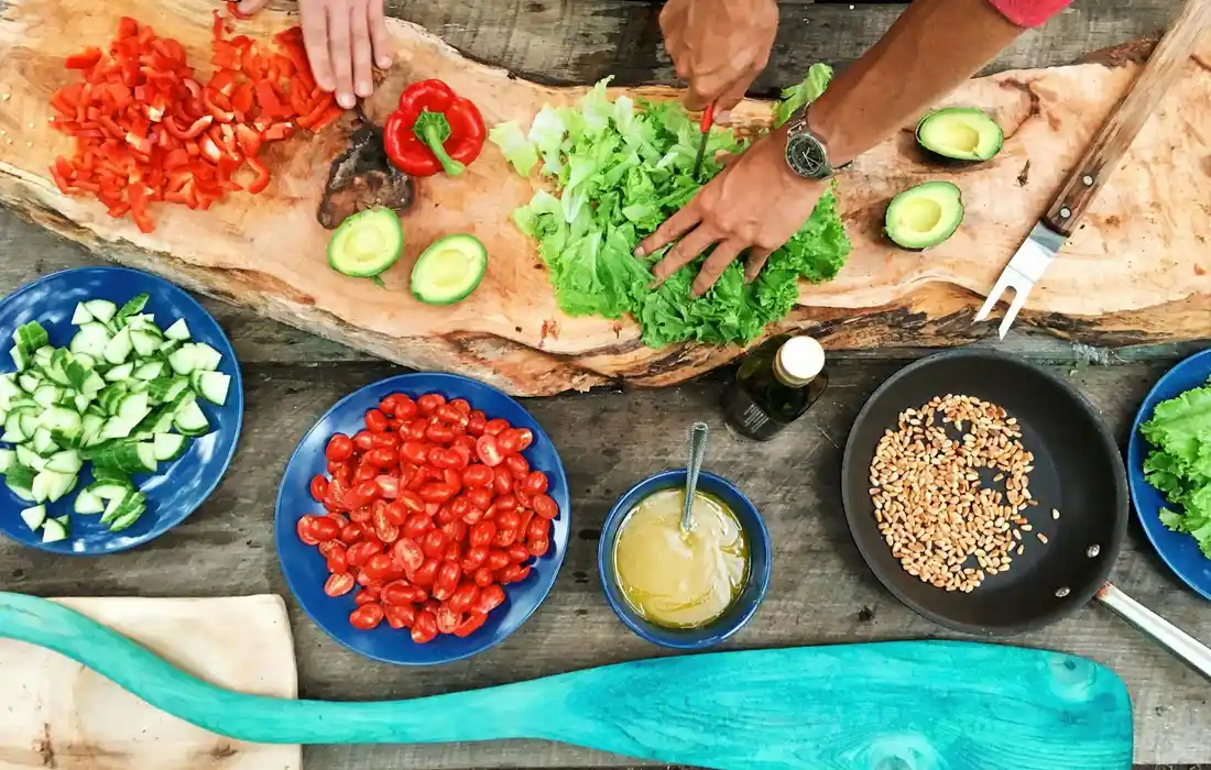 Hands chopping lettuce on a wooden board surrounded by bowls of sliced cucumber, cherry tomatoes, red peppers, and greens.