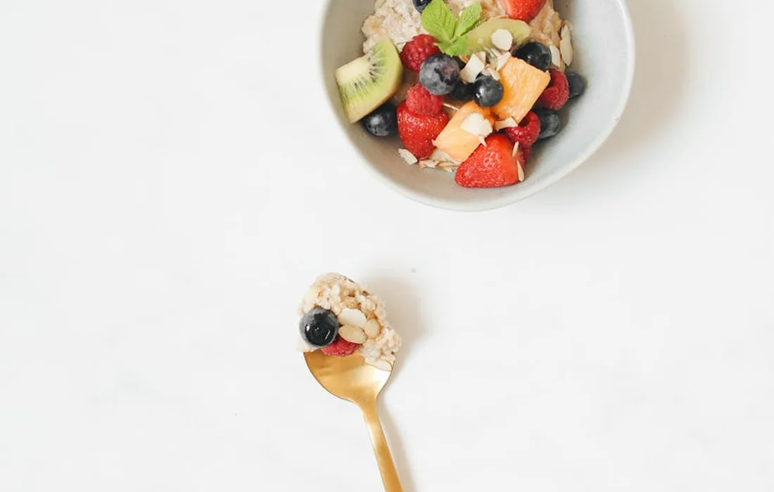 Spoonful of oats and berries held above a white surface with a bowl of mixed fruit (strawberries, blueberries, kiwi, melon) in the background