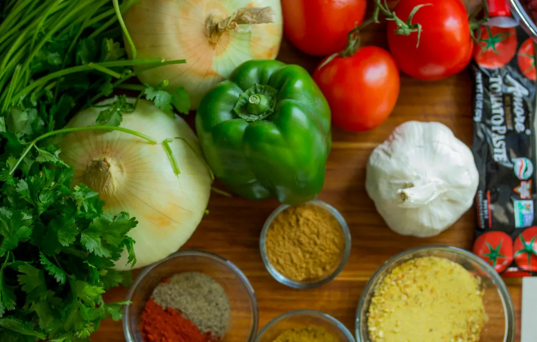 Fresh dill among other herbs and vegetables on a wooden surface, including onions, green bell pepper, garlic, tomatoes, parsley, and small bowls of spices.