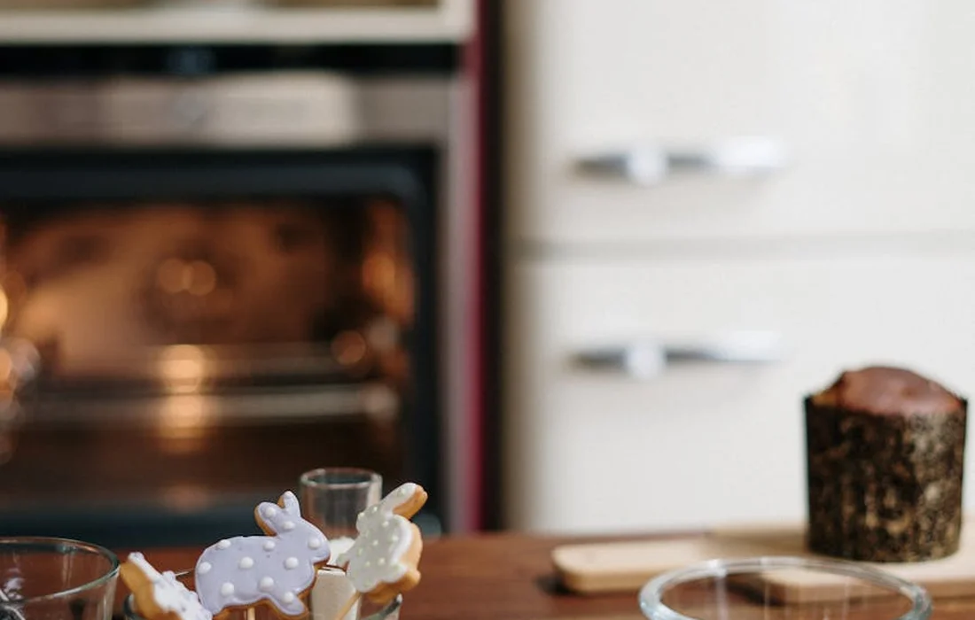 Rabbit-shaped cookies on sticks on a wooden kitchen counter with a loaf in the blurred background.