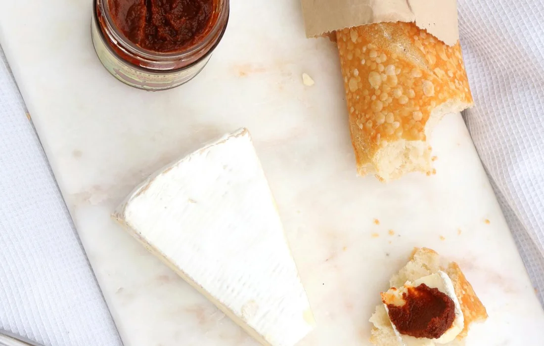 White cheese wedge, crackers, and a jar of red jam on a marble surface