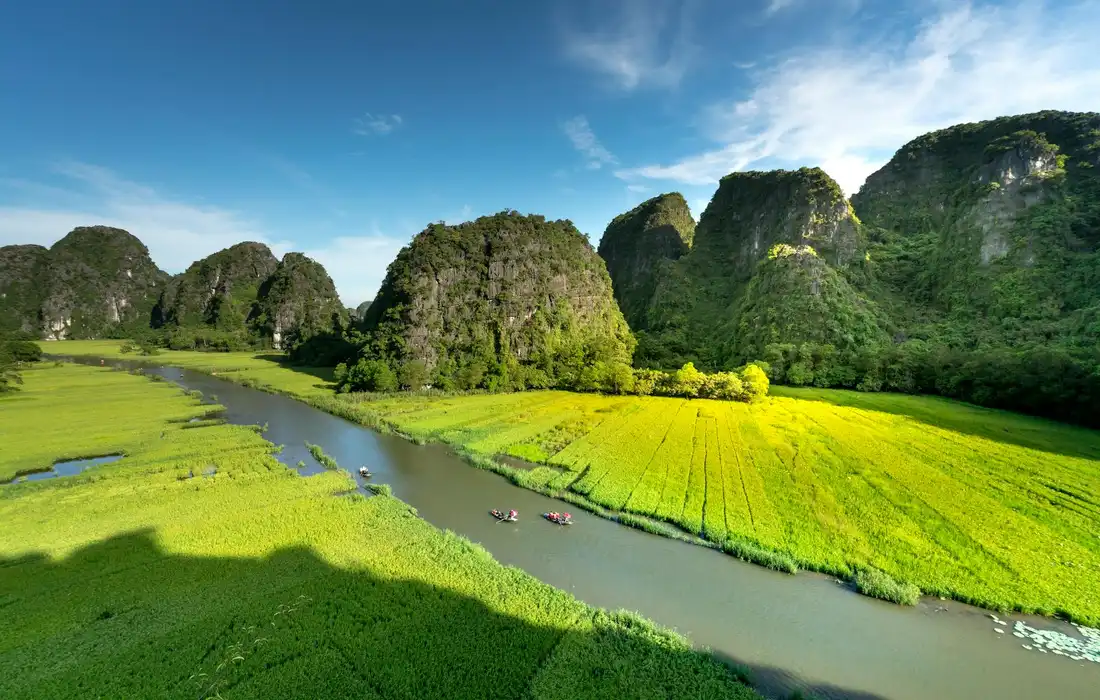Lush green fields with a winding river and limestone karst formations under a bright blue sky.