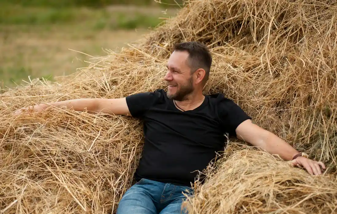A man wearing a black T-shirt and blue jeans sits among stacked hay bales outdoors, smiling.
