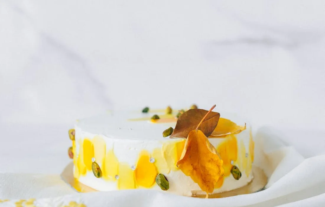 Round white-frosted cake with mango glaze dripping down the sides and pumpkin seeds on top, set on a light surface.