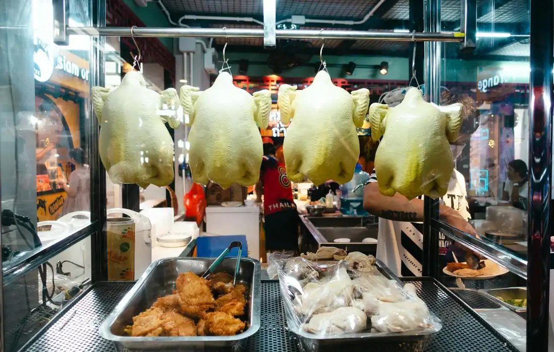 Rows of whole chickens hanging in a market stall with trays of fried items and packaged goods visible beneath.