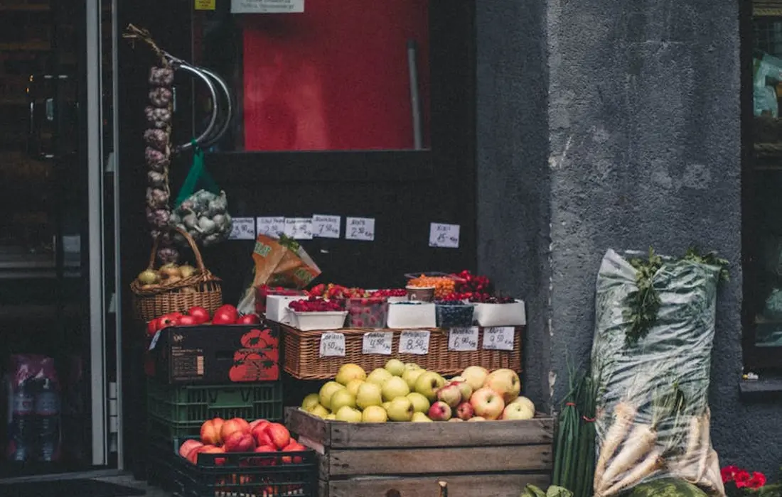 Street market stall displaying baskets of apples and pears with other produce, including leafy greens wrapped in plastic on the right.