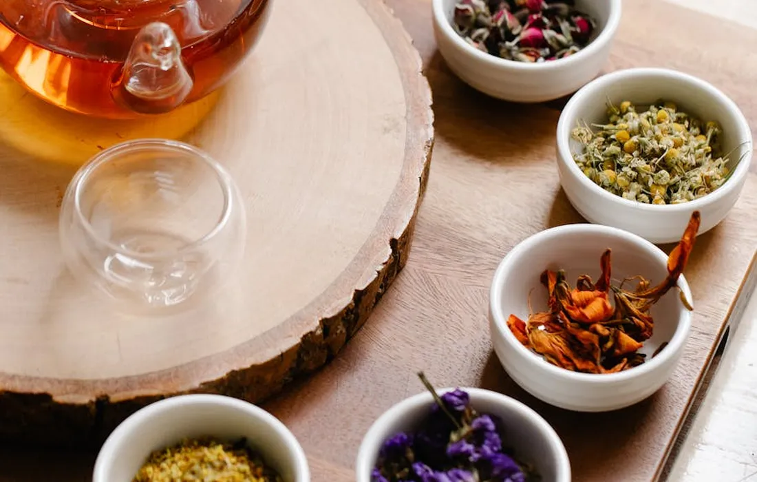 Small bowls of dried herbs including mint and dill arranged on a wooden surface beside a glass teapot.
