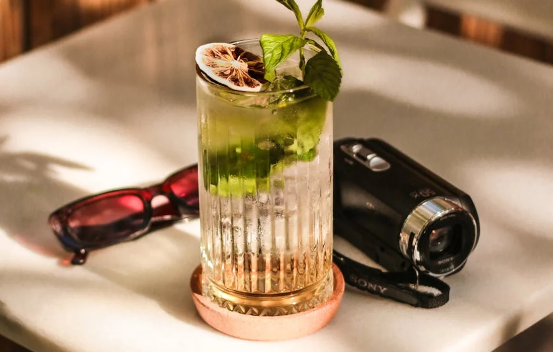 A tall glass of mint-infused beverage with fresh mint sprigs and a dried lime wheel on the rim, on a light table with sunglasses and a camera in the background.