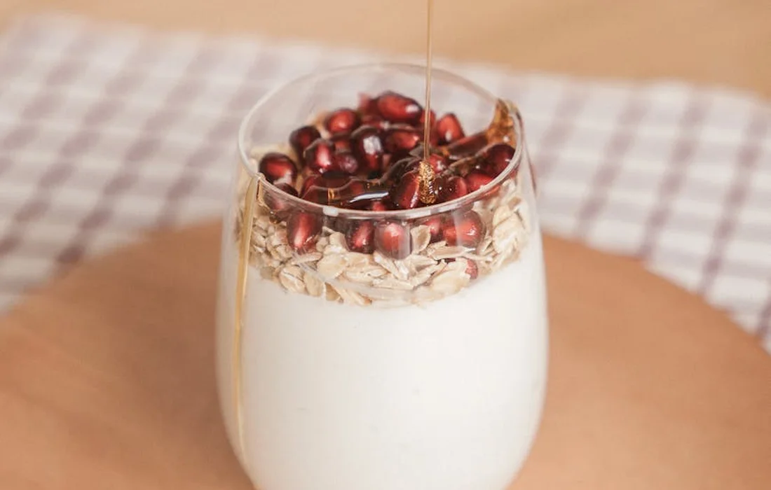 A glass with milk and layered oats, topped with red pomegranate seeds on a checkered tablecloth.