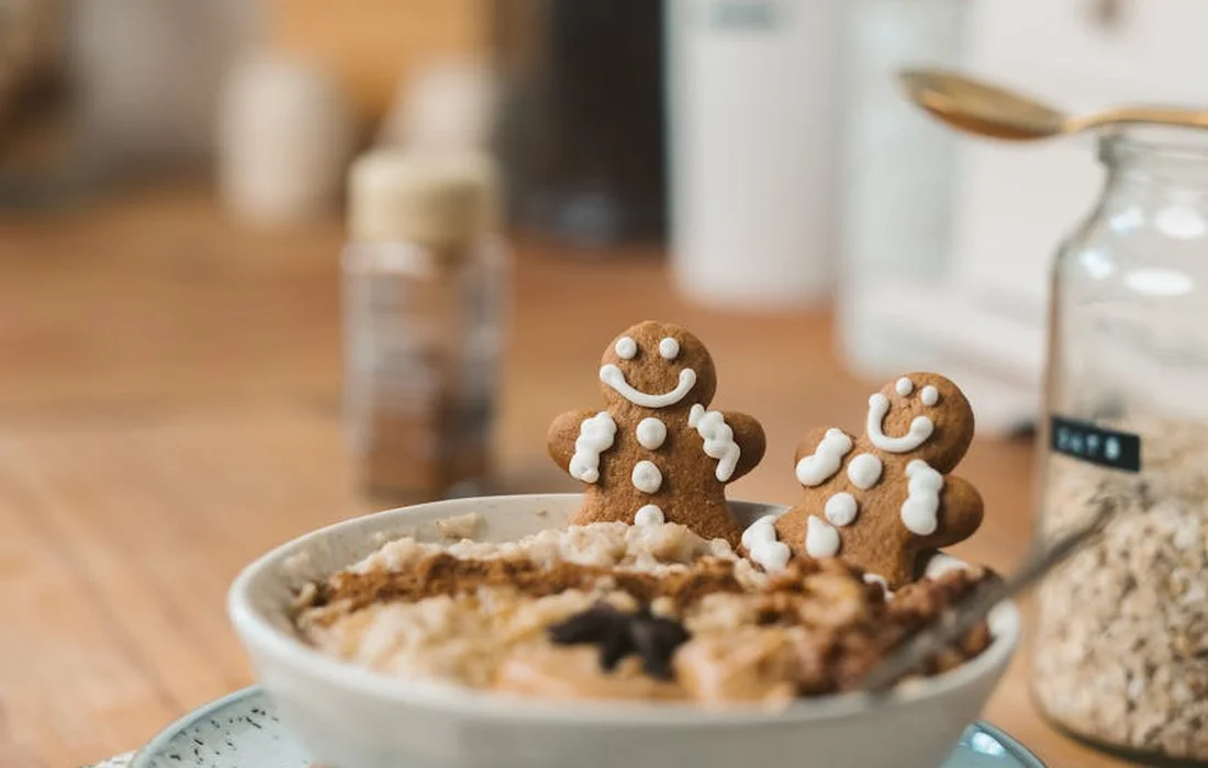A bowl of oatmeal with two gingerbread man cookies standing upright on the edge, on a wooden kitchen table with jars in the background.