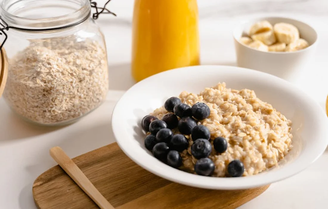 Close-up of a bowl of oats topped with blueberries on a wooden board, with a jar of oats and a bottle in the blurred background