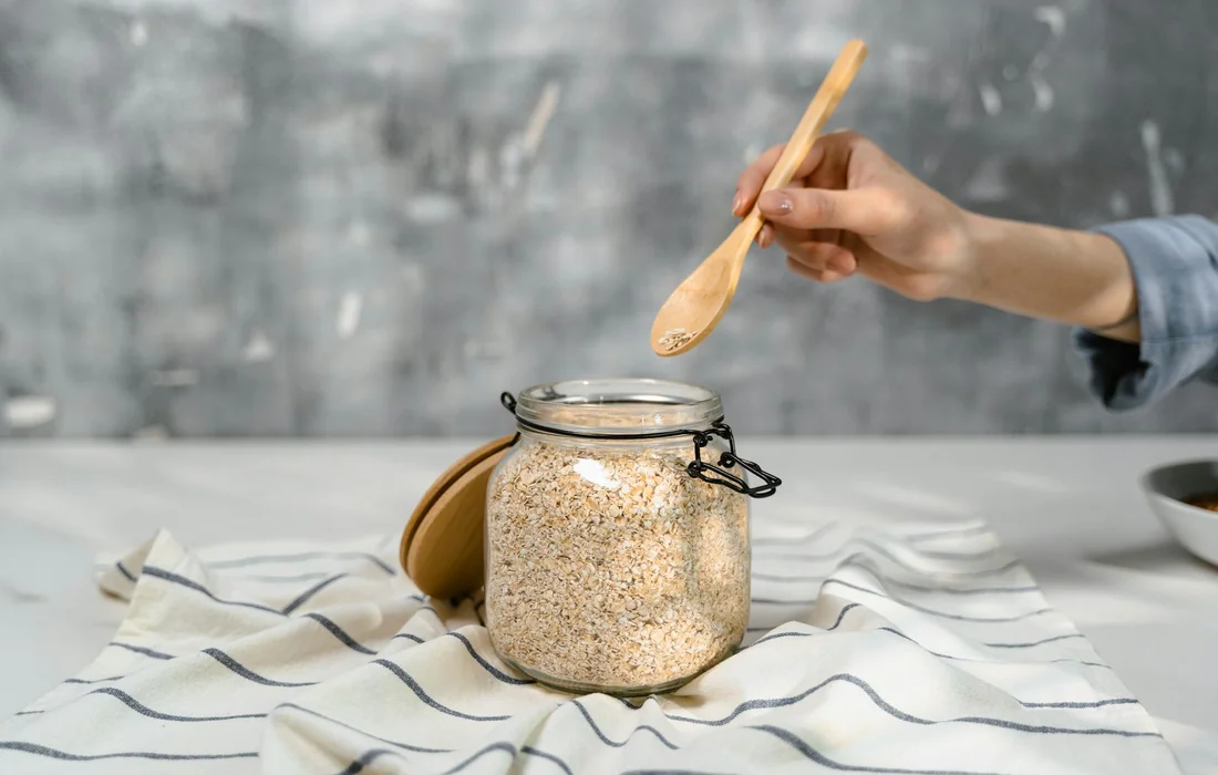 A jar of oats with a wooden spoon hovering above, set on a striped cloth in a bright kitchen.