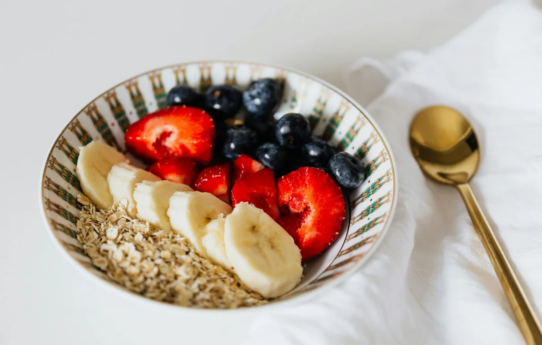 A bowl of oatmeal topped with banana slices, strawberries and blueberries, and a sprinkle of granola on a white surface beside a gold spoon