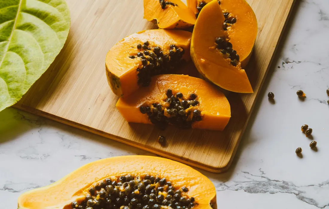 Fresh papaya slices with seeds on a wooden cutting board and a few green leaves nearby.