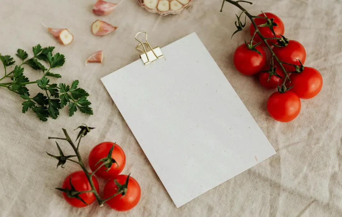 Fresh parsley sprigs with garlic and cherry tomatoes around a blank card on a beige fabric background.