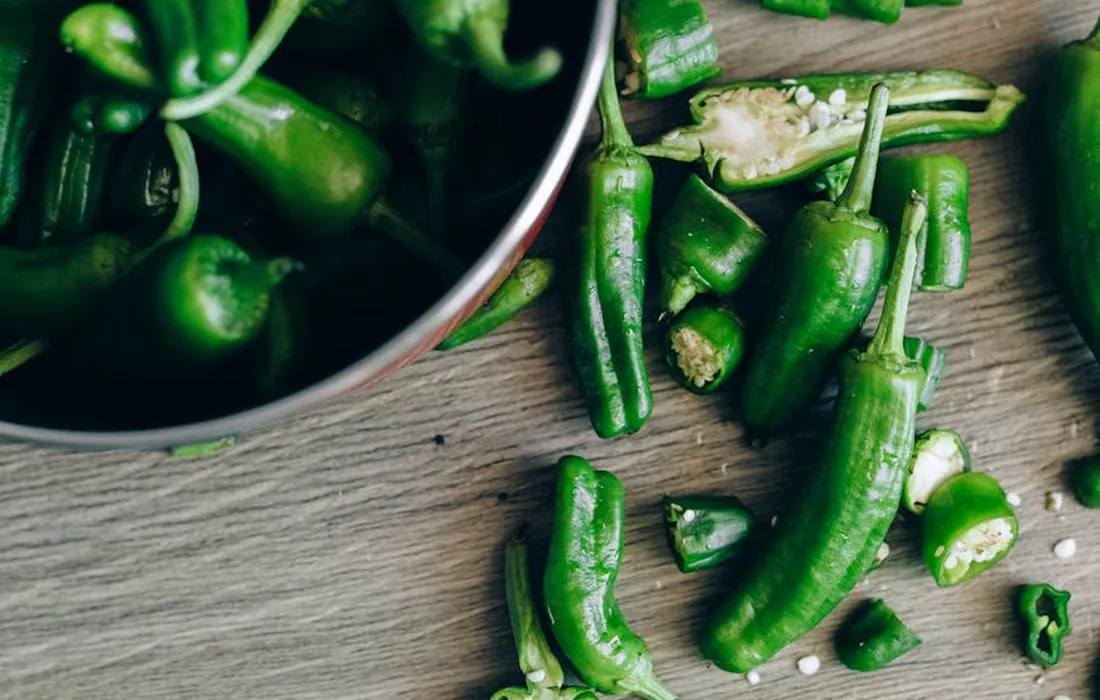Fresh green bell peppers with chopped pieces scattered on a wooden surface, illustrating pepper scraps that could be fed to other barnyard animals.