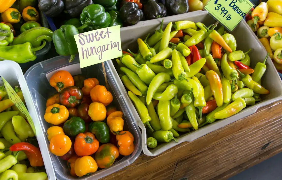 Colorful peppers in wooden crates at a market, including green chili peppers and small orange bell peppers labeled 'sweet hungarian'.