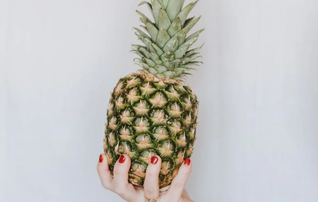 A pineapple being held by a hand with red nail polish against a pale background.