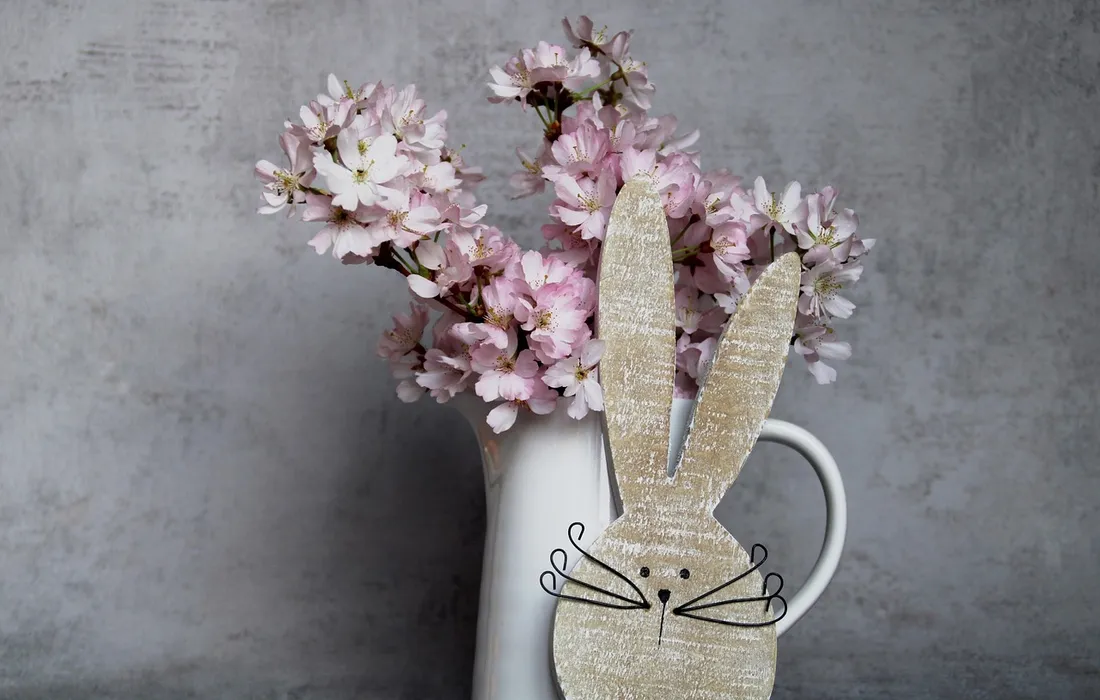 White pitcher with pink blossoms and a beige paper bunny cutout in front against a gray background.