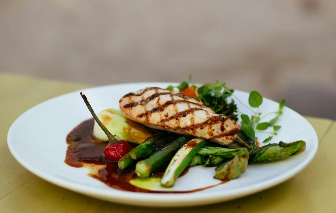 Grilled chicken breast on a bed of asparagus and other vegetables with a dark sauce on a white plate