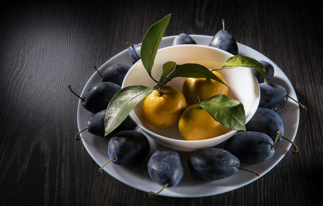 A white plate on a dark wooden surface holds a small white bowl with two yellow fruits and green leaves; the plate is surrounded by blue-purple plums.