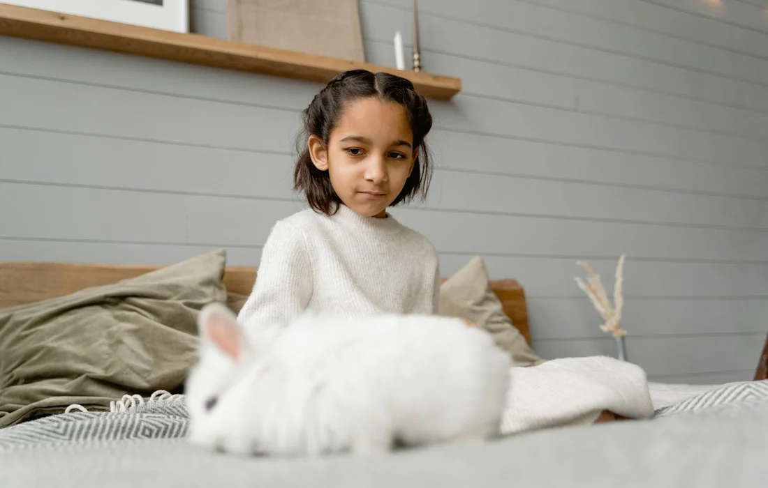 A young child sits on a bed beside a white rabbit indoors.