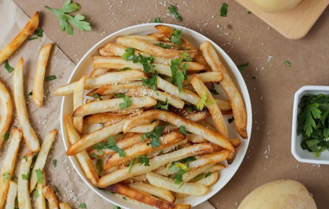 Bowl of golden potato fries garnished with parsley on a wooden surface, with extra fries and potatoes in the background.