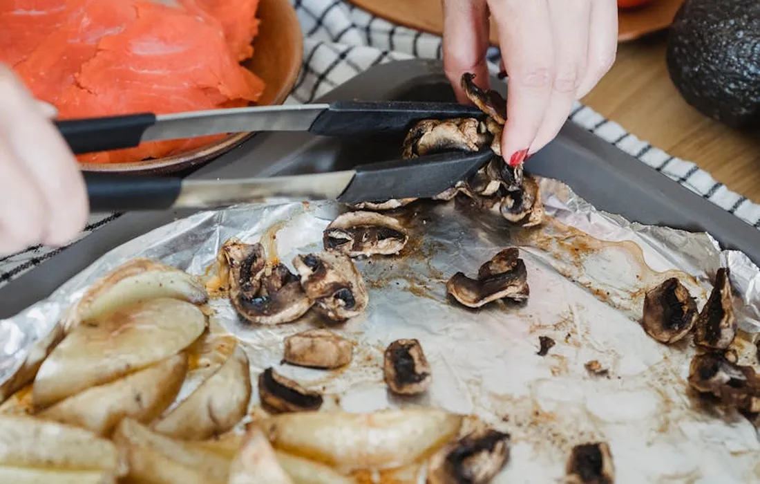Close-up of a foil-lined baking tray with sliced potatoes and mushrooms, while a hand uses tongs to move pieces.
