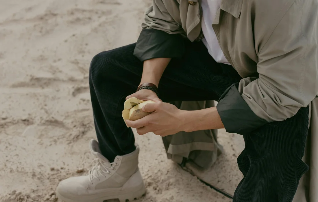 Person crouching on a sandy surface, peeling a potato outdoors, wearing a beige jacket and white sneakers.
