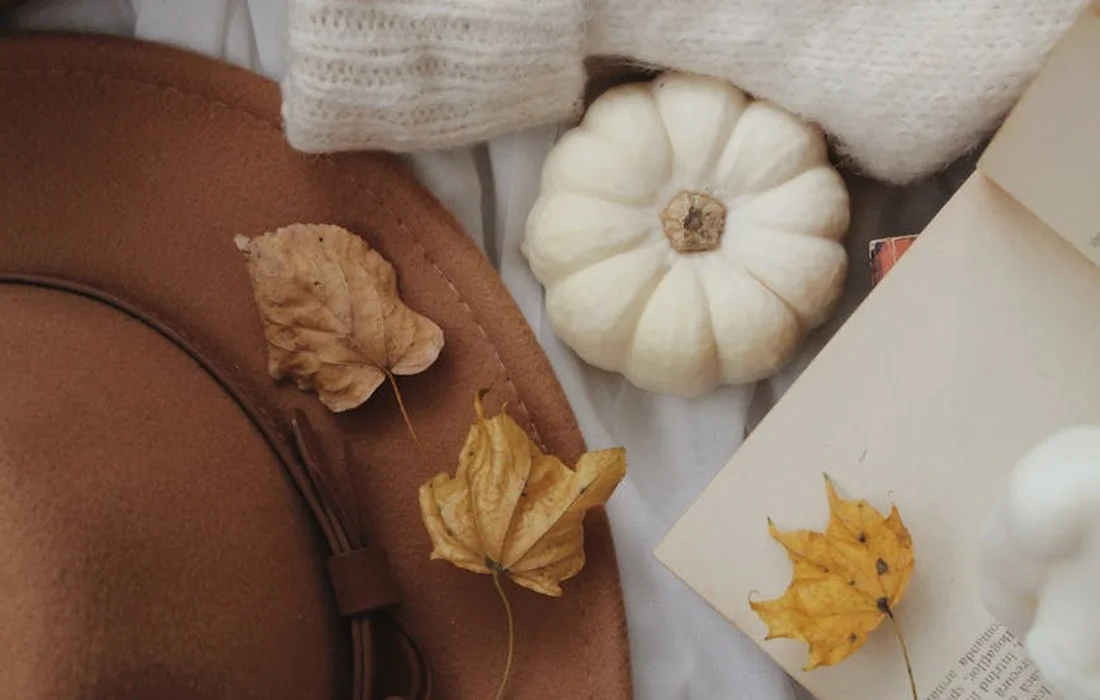 Fall flat lay featuring a small white pumpkin, dried autumn leaves, a brown hat, a book, and a cream knit blanket.