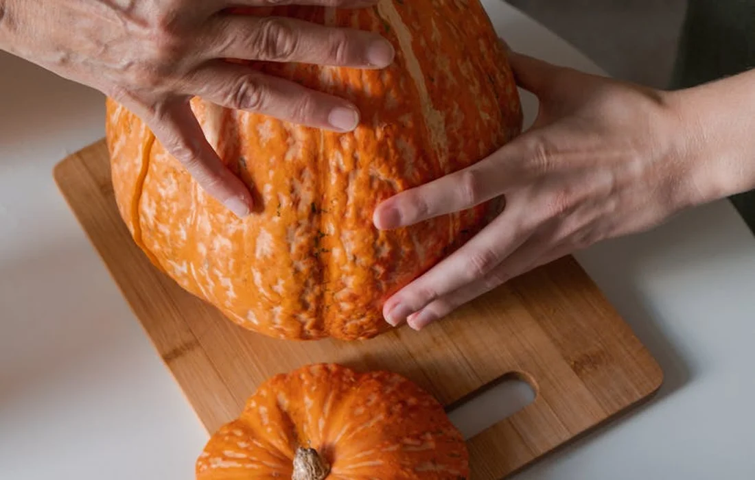 Hands cutting an orange pumpkin on a wooden cutting board