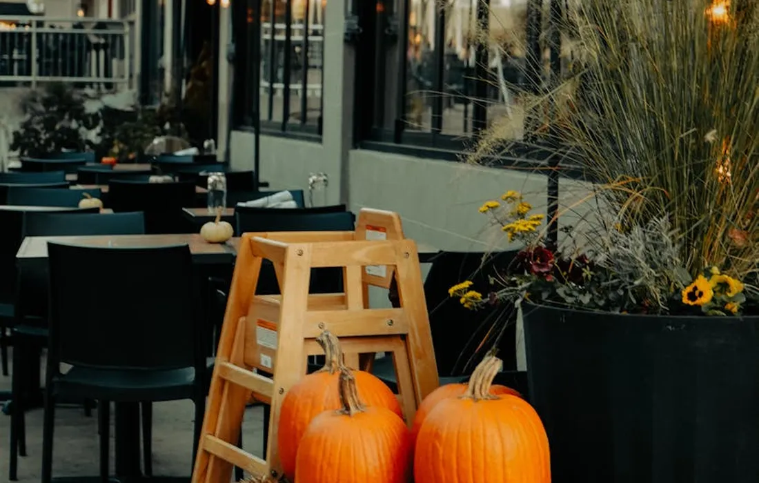 Outdoor cafe scene with bright orange pumpkins near a wooden step ladder, tables, chairs, and planters with flowers in the background.