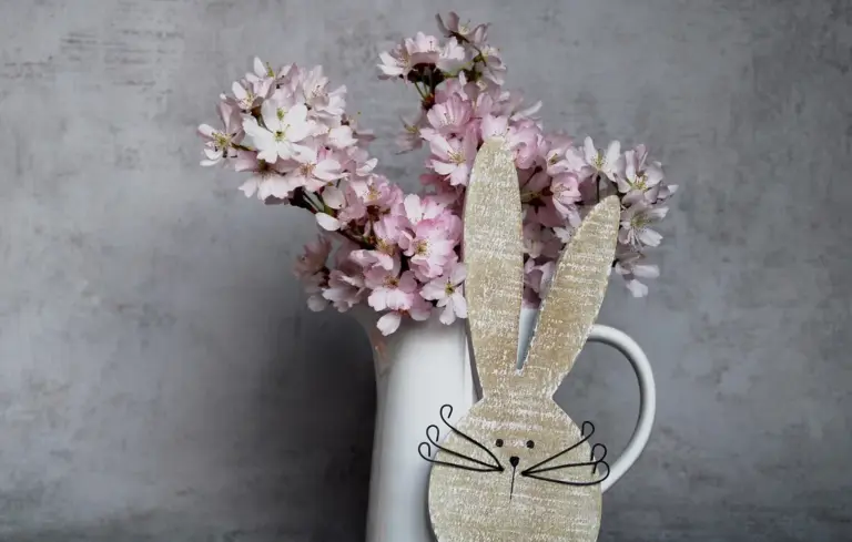 Decorative burlap rabbit cutout beside a vase of pink blossoms in a white pitcher.