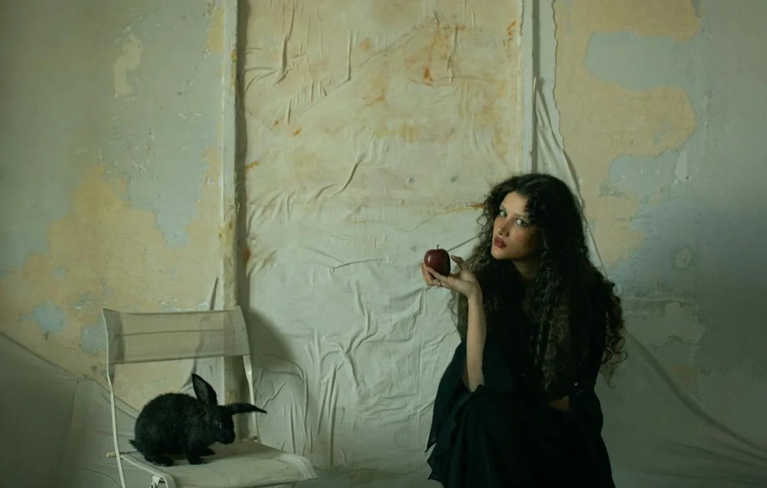 A woman with long dark hair wearing a dark dress sits beside a rabbit on a white chair in a sparse, peeling-walled room, holding an apple and looking contemplative.