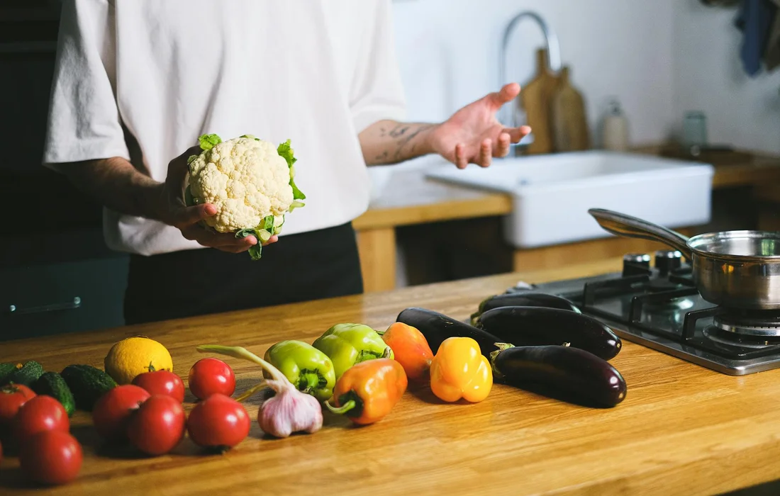Person in a white shirt holds a cauliflower while preparing fresh vegetables on a kitchen counter, including tomatoes, peppers, garlic, and eggplants.