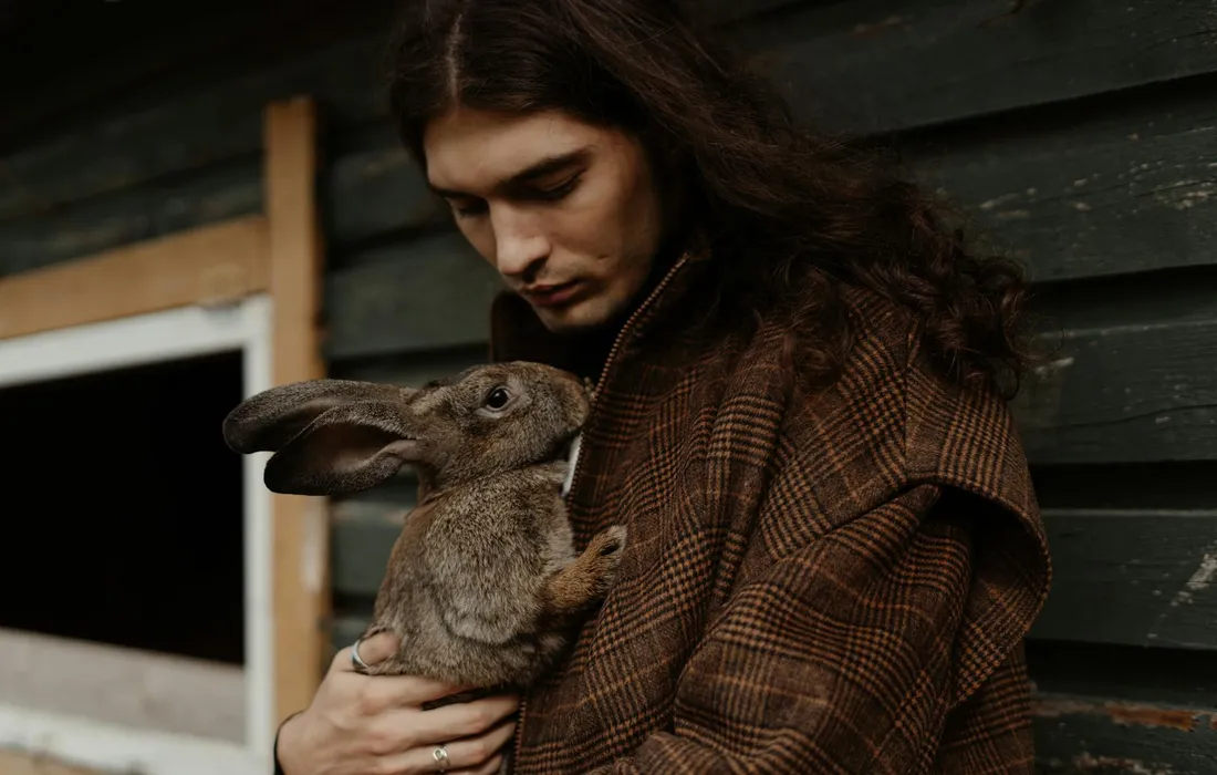 Man with long hair wearing a brown plaid coat holds a rabbit beside a green wooden wall.