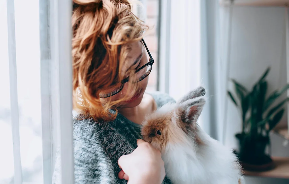 A person with short hair and glasses cradles a small fluffy rabbit by a window, looking concerned about a possible overeaten snack.