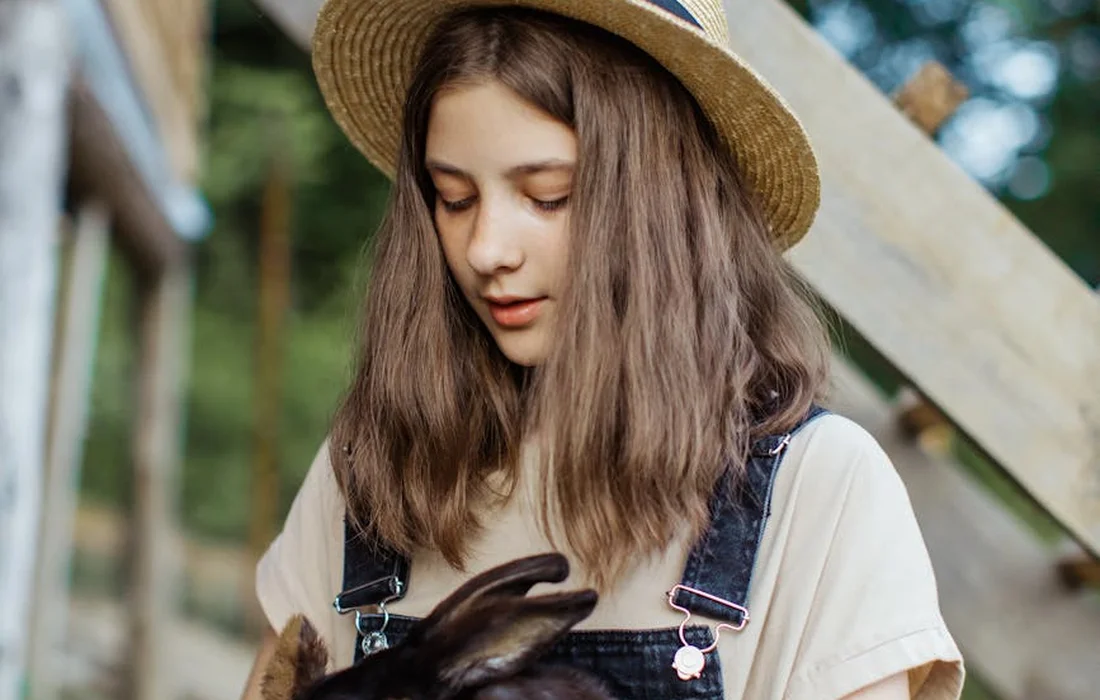 A person with long brown hair wearing a straw hat and light-colored shirt holds a rabbit outdoors.