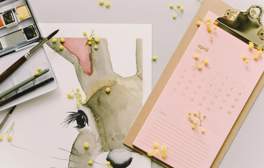 Watercolor illustration of a rabbit's head on a desk, surrounded by art supplies and a pink April calendar.