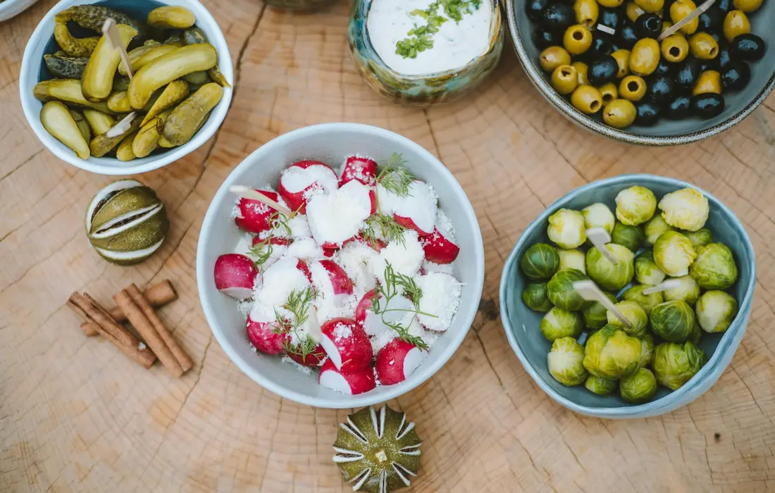 A wooden table top with bowls of fresh vegetables and herbs: center bowl of radishes with dill, left bowl of pickles, right bowl of olives, and a bowl of Brussels sprouts; cinnamon sticks lie nearby.