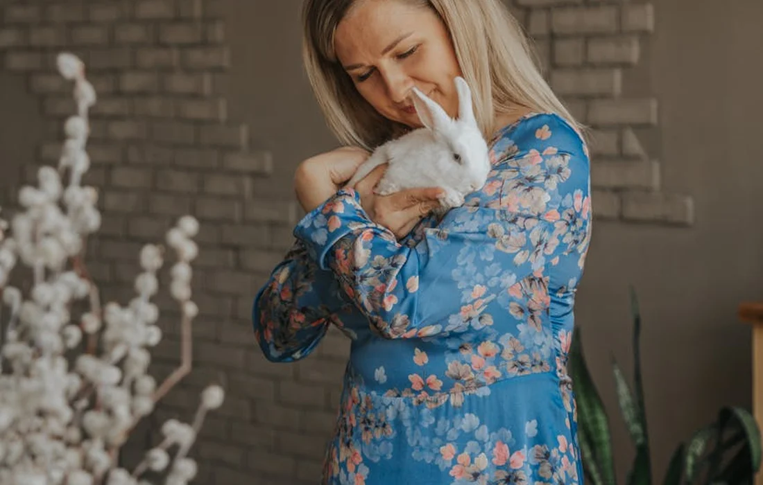 Woman in a blue floral dress cradling a white rabbit indoors.