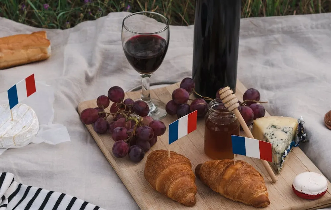 Picnic-style spread featuring grapes on a board with a wine bottle and glass, cheese, croissants, honey, and small French flags on toothpicks.