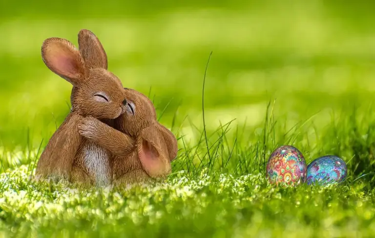 Two brown rabbits embracing in a sunlit green meadow with two colorful, decorated eggs nearby