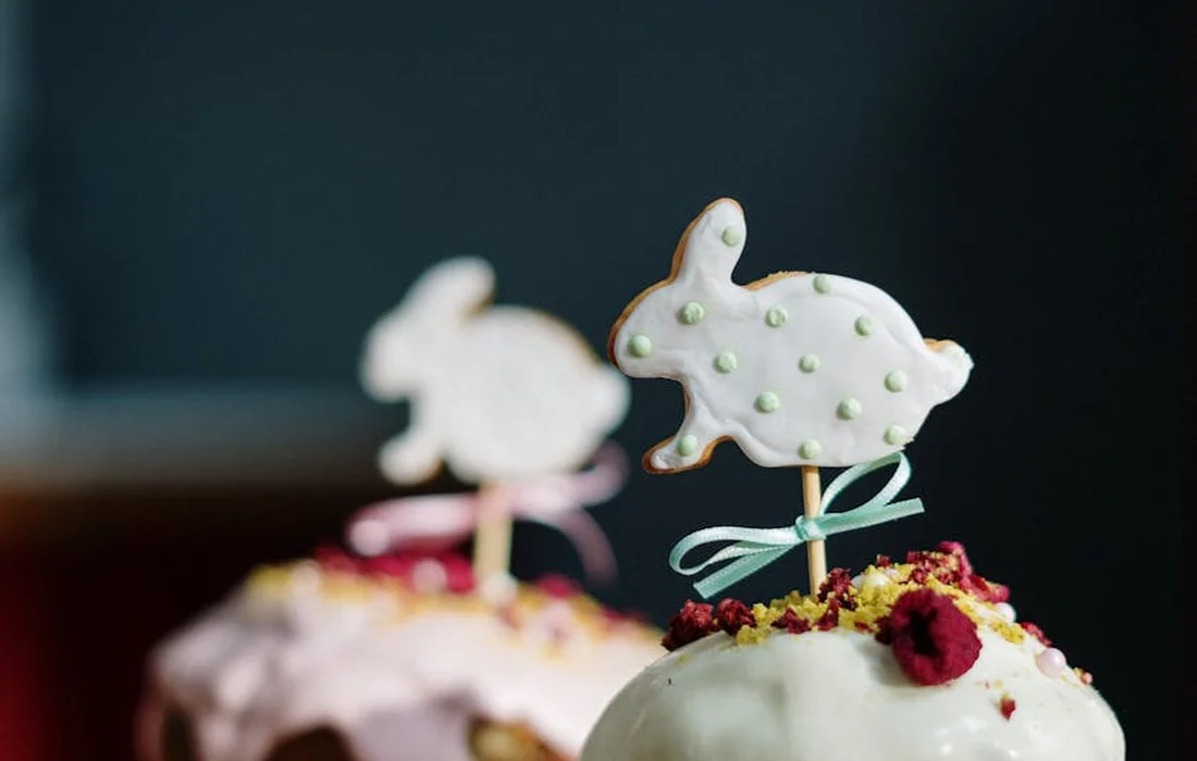 Two cupcakes topped with rabbit-shaped cookies on sticks, with pastel frosting and sprinkles, blurred background.