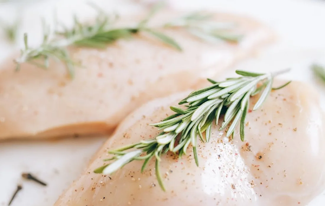 Two raw chicken breasts with a sprig of rosemary on a white surface