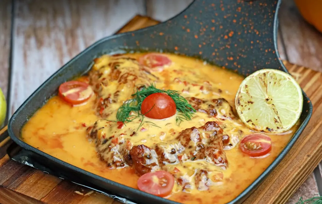 Skillet with melted cheese, tomato halves, herbs, and a lemon wedge; a tomato-based dish illustrating ripe tomatoes.