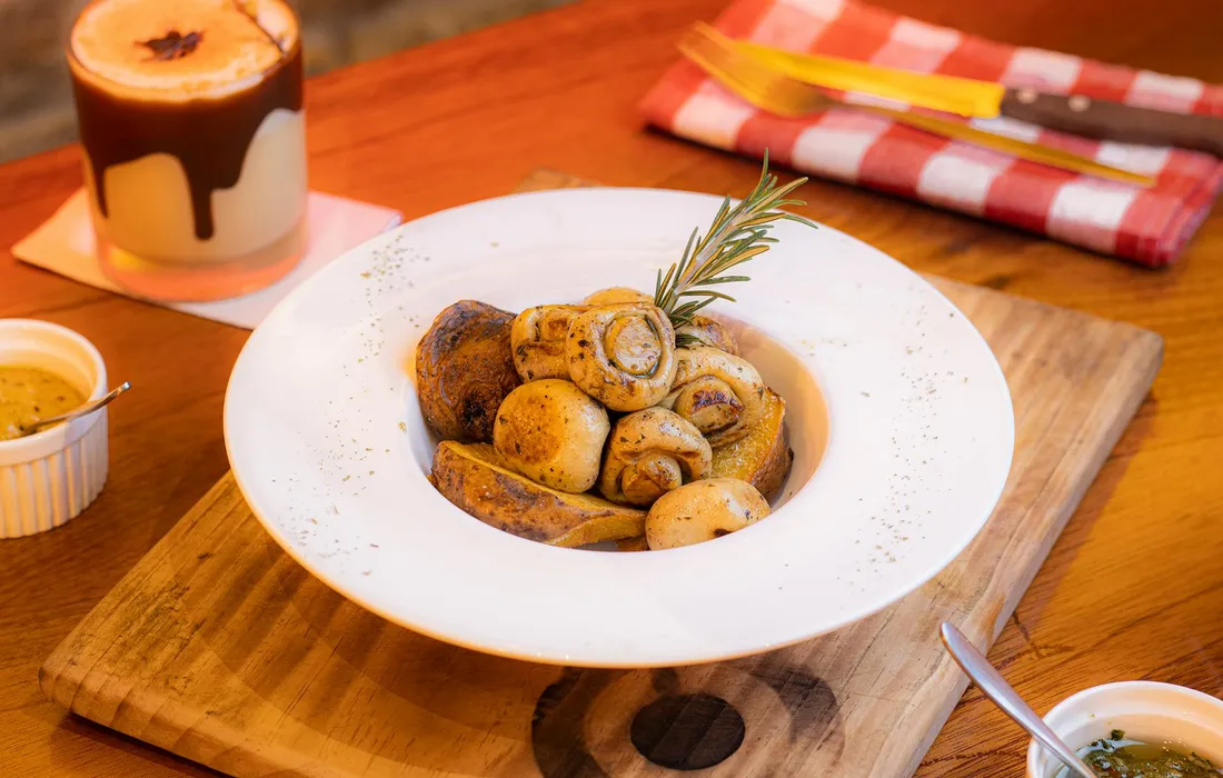 A white bowl of roasted baby potatoes garnished with a rosemary sprig, placed on a wooden board with a checkered napkin and candle in the background.